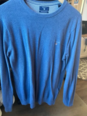 Blå stickad tröja från Gant - Snygg blå stickad tröja från Gant i premium bomull. Tröjan har rund halsringning, diskret Gant-logga broderad på bröstet och ribbade muddar vid ärmslut och nederkant. Perfekt för lager på lager och enkel att matcha med jeans eller chinos.