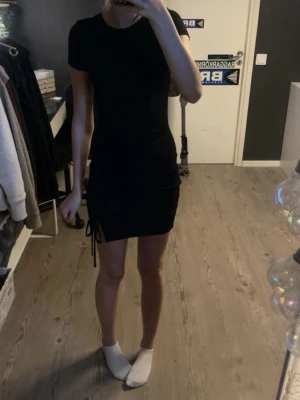 Svart kort klänning med snörning - Säljer en svart klänning från H&M med snörning längs sidan som ger en snygg rynkad effekt. Klänningen har korta ärmar. Enkel och trendig modell.  Helt oanvänd med prislapp kvar 💗