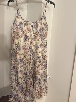 Blommig sommarklänning med volanger - Söt blommig klänning i ljusa toner med volangdetaljer och smala axelband. Klänningen har ett romantiskt mönster i lila, rosa och grönt på vit botten. Perfekt för varma dagar och har en luftig, flowy passform 