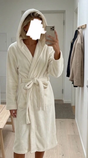 Krämvit fluffig morgonrock med huva - Mysig krämvit morgonrock i mjuk fleece med huva och knytskärp i midjan. Långa ärmar och två stora fickor framtill. Perfekt för att chilla hemma och hålla sig varm under kalla morgnar.