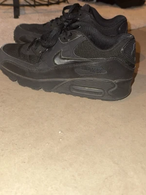 Svarta Nike Air Max 90 sneakers - Snygga helsvarta Nike Air Max 90 sneakers med klassisk siluett. Skorna har ovandel i mesh och läder, synlig Air-enhet i sulan och ikonisk Swoosh på sidan. Perfekta för dig som gillar streetwear och vill ha en tidlös look.