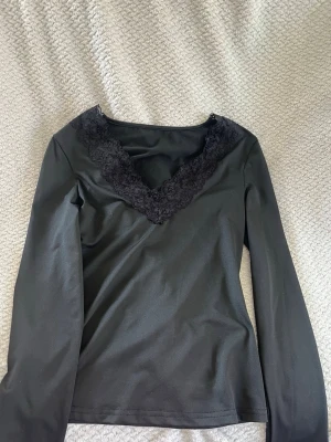 Svart långärmad topp med spets - Snygg svart långärmad topp med v-ringning dekorerad med bred spetskant. Toppen har en figurnära passform och är perfekt för att lyfta vilken outfit som helst. Tillverkad i mjukt material som känns skönt mot huden.
