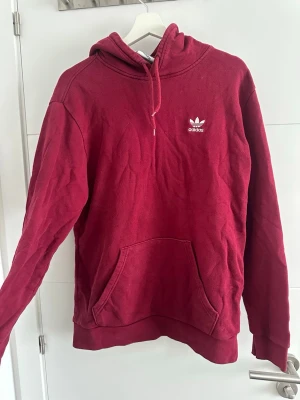 Vinröd hoodie från Adidas - Säljer en vinröd hoodie från Adidas med klassisk luva och magficka. Tröjan har Adidas logga broderad i vitt på bröstet och ribbade muddar vid ärmslut och nederkant. Perfekt för en avslappnad och sportig stil.