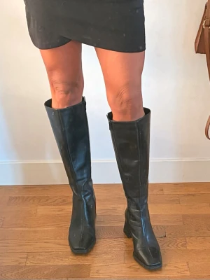 Svarta knähöga boots med klack - Snygga svarta knähöga boots i skinn med fyrkantig tå och bred klack. Dragkedja på insidan för enkel påtagning. Perfekta till kjol eller klänning för en trendig look. Vagabond Hedda. Nypris 2500 kr.