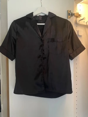 Svart kortärmad satinpyjamas - Stilren svart pyjamasskjorta och shorts i glansigt satinmaterial. Skjortan har knäppning framtill och en bröstficka för extra detalj. 