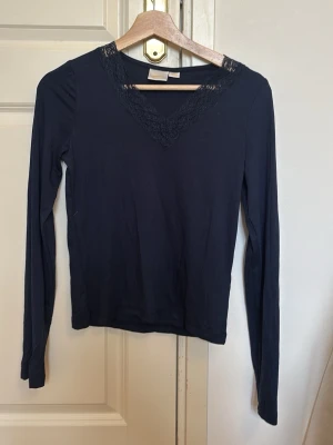 Svart långärmad topp med spets - Snygg svart långärmad topp med v-ringning och spetsdetalj vid halsen. Perfekt att matcha med jeans eller kjol för en stilren look. Materialet känns mjukt och stretchigt, vilket gör den bekväm att bära hela dagen.