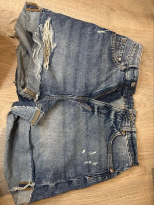 Blå jeansshorts - Säljer ett par blå jeansshorts med slitningar och råa kanter. Shortsen har klassisk femficksdesign, dragkedja och knapp framtill samt uppvikta benslut. Perfekta för en avslappnad och trendig sommarlook. Använda fåtal gånger men är i perfekt skick. 