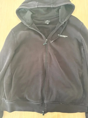 Svart hoodie från Armani Exchange - Svart hoodie med dragkedja från Armani Exchange. Klassisk modell med huva och fickor på sidorna. Diskret logga på bröstet. Tillverkad i mjukt material som känns skönt mot huden. Perfekt för en avslappnad och stilren look.