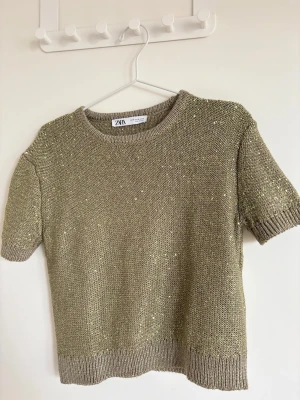 Glittrig beige stickad topp från ZARA - Säljer en glittrig beige stickad topp från ZARA med korta ärmar och rund hals. Toppen har insydda små paljetter som ger ett snyggt skimmer och ribbade kanter vid ärmslut och nederkant. Perfekt för dig som vill ha något extra till din outfit.