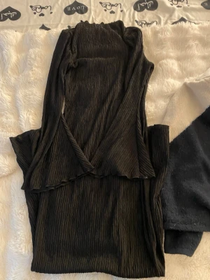Svart omlottklänning med volang - Säljer en svart omlottklänning med volangdetaljer och ribbad struktur. Klänningen har en smickrande v-ringning och är tillverkad i ett mjukt, stretchigt material. Perfekt för dig som gillar en stilren och trendig look.