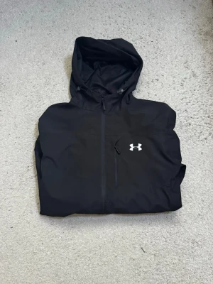 Svart vindjacka från Under Armour - Snygg svart vindjacka från Under Armour med huva och dragkedja framtill. Jackan har en vit logga på bröstet och praktiska fickor med dragkedja. Perfekt för blåsiga dagar och sportiga outfits.