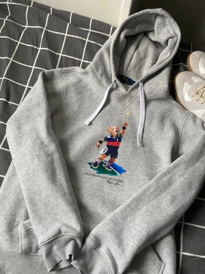 Grå hoodie Polo Bear Ralph Lauren - Säljer en grå hoodie från Polo Ralph Lauren med den ikoniska Polo Bear-figuren på bröstet. Hoodien har huva med vita snören, ribbade muddar och en klassisk känguruficka. Perfekt för dig som gillar streetwear och vill ha en stilren men lekfull look.