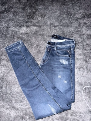 Blå slitna slimfit jeans Replay - Snygga blå slim fit  jeans från Replay med slitningar på framsidan och klassisk femficksdesign. Jeansen har en smal passform och är tillverkade i stretchigt denimtyg för extra komfort. Perfekta för en trendig och avslappnad look.