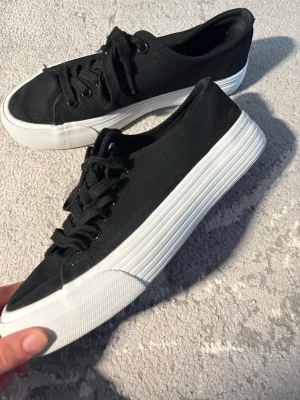 Svarta låga sneakers från Anna Field  - Snygga svarta sneakers från Mansfield med klassisk låg profil och vit tjock sula. Skorna har svarta skosnören och är tillverkade i canvasmaterial som ger en avslappnad vibe. Perfekta för dig som gillar enkel och stilren design.