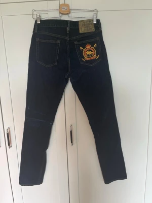 Jeans Ralph Lauren - Snygga mörkblå jeans i slim fit och klassisk femficksmodell. Ena bakfickan har en stor broderad patch i gult och rött med krona och emblem. Jeansen har kontrastsömmar och gylf med knapp. Perfekta för dig som vill sticka ut lite extra.