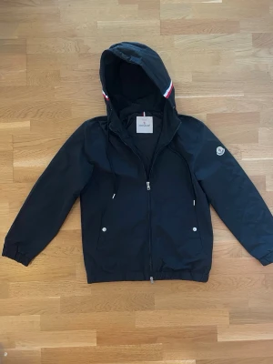 Svart vindjacka från Moncler - Snygg svart vindjacka från Moncler med huva och dragkedja framtill. Jackan har två fickor med tryckknappar och Moncler-logga på ärmen. Detaljer i rött och vitt på huvan ger en sportig touch. Perfekt för blåsiga dagar. Står M men passar som S.