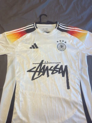 Vit T-shirt Adidas x Stüssy DFB - Säljer en vit T-shirt från Adidas i samarbete med Stüssy och tyska landslaget. Tröjan har korta ärmar, svarta Adidas-ränder på axlarna och färgdetaljer i rött, orange och gult. Stüssy-logga tryckt framtill och DFB-märke med fyra stjärnor på bröstet.
