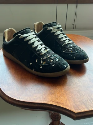Svarta sneakers med färgstänk - Unika svarta sneakers med vita snören och beige insida. Skorna har coola färgstänk i vitt, gult och blått på tåpartiet som ger en kreativ vibe. Yttermaterialet är en mix av mocka och läder, och sulan är beige. Perfekta för dig som vill sticka ut.