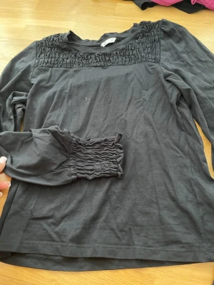 Svart långärmad topp med rynk - Svart långärmad topp med rynkdetaljer över bröstet och vid ärmsluten. Toppen har en enkel rund halsringning och är gjord i mjukt material som känns skönt mot huden. Perfekt för dig som gillar stilrena plagg med en twist.