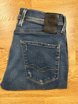 Blå jeans från Replay hyperflex  - Snygga blå jeans från Replay med klassisk femficksmodell och slitna detaljer. Tillverkade i ett mjukt denimtyg som sitter skönt och har en modern passform. De är hyperflex vilket gör att dem sitter ännu skjönare än vanliga jeans som inte har de.