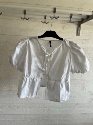 Vit blus med puffärm och knyt - Söt vit blus i storlek XS. Blusen har korta puffärmar, v-ringning och knyt framtill. Tillverkad i ett lätt tyg som ger en luftig känsla. Perfekt till jeans eller kjol för en trendig look.