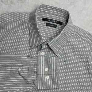 Tiger of Sweden N. Randy ST 140's Double skjorta randig grå/vit storlek 40 / L - Randig skjorta från Tiger of Sweden i modellen N. Randy ST. Klassisk grå och vit smalrandig design som passar lika bra till kostym som till chinos eller jeans. Tillverkad i exklusiv 140's Double-bomull för en tunn, mjuk och följsam känsla. Modellen är tillverkad i 100% bomull och producerad i Vietnam.   Storlek 40 / L  Mycket fint skick.  Mått Bröstbredd: ca 50 cm Plagglängd: ca 70 cm Axelbredd: ca 44 cm Ärmlängd: ca 67 cm  Material 100% bomull  Klassisk krage och smalrandigt mönster.