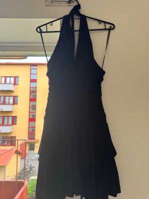 Svart kort halterneckklänning - Snygg svart kort klänning med halterneck och volangdetaljer nertill. Klänningen har en omlottkänsla och knyts i nacken. Perfekt för dig som vill ha en stilren och trendig look.