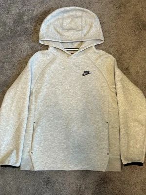 Ljusgrå hoodie från Nike - Snygg ljusgrå hoodie från Nike med svart logga på bröstet. Hoodien har en klassisk huva och raka ärmar med svarta detaljer vid ärmsluten. Tillverkad i mjukt material som känns skönt mot huden. Perfekt för en avslappnad och sportig stil.