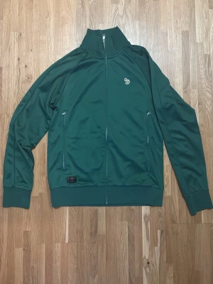 Mörkgrön Paul Smith ziptröja med hög krage - Snygg mörkgrön ziptröja med hög krage och två fickor med dragkedja framtill. Tröjan har ribbade muddar vid ärmslut och nederkant samt en liten broderad logga på bröstet. Perfekt för en sportig och avslappnad stil.