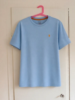 Polo Ralph Lauren T-shirt i ljusblått 🩵 - Klassisk modell med orange broderad Polo-logga på bröstet och orange detalj vid insidan av halsen. Enkel och snygg färgkombination som passar perfekt till sommaren. Storlek L (14–16), ca 160, junior. Fint begagnat skick men den har några fläckar! 🏷️ Polo Ralph Lauren 📏 L (14–16) / 160 🎨 Ljusblå med orange detaljer