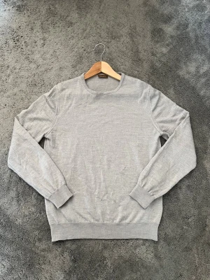Crew Neck Merinoull Tröja - En rikigt kvalité och stilren ljusgrå tröja med rund halsringning och långa ärmar. Tröjan har ribbade muddar vid ärmslut och nederkant. Perfekt att matcha med jeans nu till hösten. Mycket bra skick på plagget i sig, använd endast ett fåtal gånger. Minimalt slitage i ena armhålan men påverkar varken looken eller användningen. Pris går att diskuteras! 