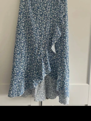 Blommig blå omlottkjol med volang - Söt blå omlottkjol med vitt blommönster och volangkant. Kjolen har en luftig känsla och passar perfekt till varma dagar. Materialet känns lätt och skönt mot huden.