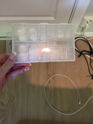 Vita press-on naglar i ask - Säljer ett set med vita press-on naglar i olika storlekar, levereras i en praktisk plastask med fack. Naglarna har en klassisk fyrkantig form och är tillverkade i plast. Perfekt för dig som vill fixa snygga naglar snabbt och enkelt.