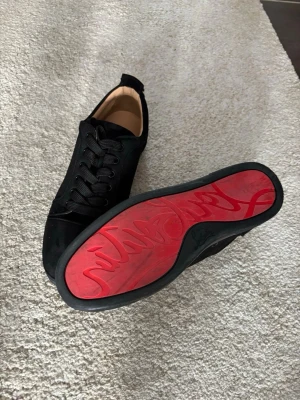Svarta Loubs - Stilrena svarta sneakers i mockaimitation med svarta skosnören och låg profil. Insidan är beige och sulan är svart med en unik röd undersida och grafiskt mönster. Perfekta för dig som vill ha en clean look med en twist.