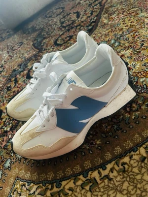 Vita New Balance sneakers med blå detalj - Snygga vita sneakers från New Balance med blå logga på sidan och beige mockadetaljer vid tån. Skorna har vita snören, mesh och lädermaterial samt svart yttersula. Perfekta för en sportig och trendig look.