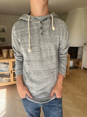 Pull & Bear henley hoodie grå herr - Gråmelerad luvtröja från Pull & Bear i mjukt material. Knappar i halsen (henley-modell) och ljusa, grova dragskor till huvan. Praktisk magficka framtill. Väldigt skön och i mycket gott skick!#pullandbear #hoodie #henley #melerad #gråtröja #luvtröja #casual #streetwear #herrtröja #menswear #pullbear #knappar #mysig #vardagsstil #fashion #sköntröja #herrkläder #vintedse #trendig