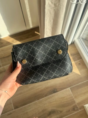 Svart axelväska med rutmönster - Snygg svart axelväska med diskret rutmönster och guldfärgade knappar framtill. Väskan har en enkel, stilren design och justerbar axelrem. Perfekt storlek för det viktigaste och passar till många olika outfits.