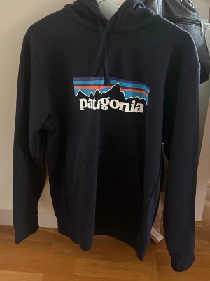 Svart Patagonia hoodie med tryck - Snygg svart hoodie från Patagonia med det klassiska logotrycket i vitt, blått, lila och orange på bröstet. Hoodien har huva med dragsko och en stor magficka. Perfekt för dig som gillar streetwear och outdoor-vibbar.
