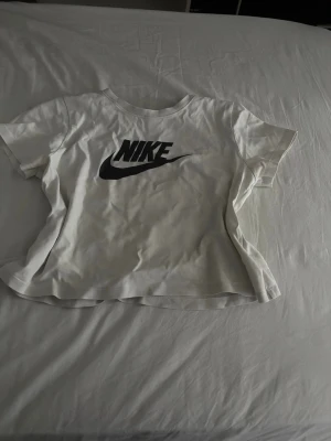 Vit Nike t-shirt med logga - Snygg vit t-shirt från Nike med klassisk svart logga på bröstet. T-shirten har rund halsringning och korta ärmar. Perfekt för en sportig och avslappnad stil. Tillverkad i mjuk bomull för skön komfort.