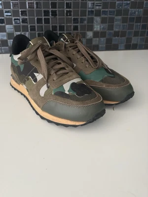 Valentino rockruners - Snygga sneakers med camouflage-mönster i grönt, brunt och svart. Skorna har mockadetaljer, gröna och beige partier samt svarta sulor. Klassisk låg modell med bruna skosnören och bekväm passform. Perfekta för dig som gillar streetwear och vill sticka ut.