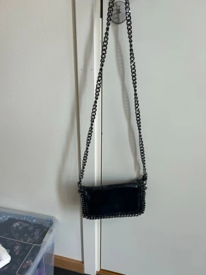 Svart axelväska med kedjerem - Snygg svart axelväska i rektangulär form med grov silverfärgad kedjerem och kedjedetaljer runt kanterna. Väskan har en stilren look och är tillverkad i ett material som ser ut som konstläder. Perfekt för dig som gillar edgy accessoarer. Köpt i Italien 