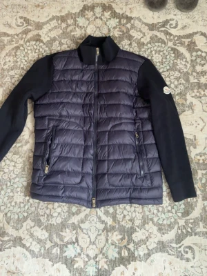 Mörkblå cardigan  från Moncler - Snygg mörkblå dunväst från Moncler med stickade ärmar och quiltad front. Två dragkedjeförsedda fickor framtill och klassisk Moncler-logga på ärmen. Perfekt för lager-på-lager och ger en sportig men stilren look.