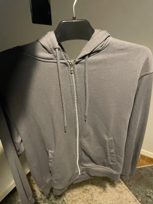 Grå zip hoodie med fickor - En stilren grå hoodie med dragkedja framtill och två fickor. Tröjan har huva med dragsko och ribbade muddar vid ärmslut och nederkant. Perfekt för en avslappnad look och enkel att matcha med andra plagg.