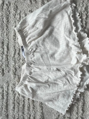Vita pyjamasshorts med volangkant - Säljer ett par vita pyjamasshorts i mjukt ribbat tyg med diskreta ränder. Shortsen har elastisk midja och söta volangkanter nedtill för en extra gullig look. Perfekta för mysiga hemmakvällar eller sommarens varma nätter.