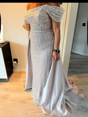 Glittrig offshoulder långklänning - Elegant glittrig långklänning i ljus champagnefärg med offshoulder och draperad överdel. Klänningen har slits och vackra paljettdetaljer som ger extra glamour. Perfekt för bal eller festliga tillfällen.