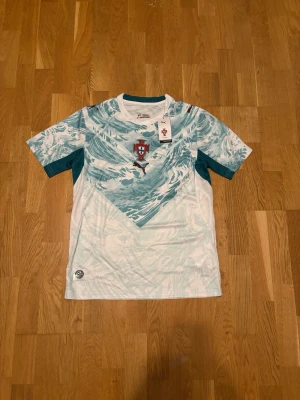 Portugal t-shirt med vågmönster Puma - Snygg t-shirt från Puma med Portugals landslagsemblem på bröstet. T-shirten har ett unikt våginspirerat mönster i ljusblått och vitt, rund halsringning och korta ärmar. Perfekt för dig som gillar fotboll och vill sticka ut.
