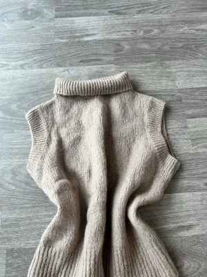 Beige stickad poloväst - Mysig stickad väst i beige med hög polokrage och ribbade kanter. Ärmlös modell som är perfekt att styla över en skjorta eller t-shirt. Materialet känns mjukt och ger en avslappnad men ändå trendig look.