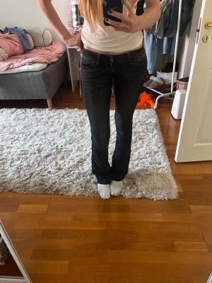 Mörk gråa bootcut jeans med låg midja - Säljer ett par mörk gråa  bootcut jeans med låg midja och små rosa detaljer detaljer på fickorna.  Jag säljer dessa för att det tyvärr är lite små i midjan men det är inte för korta på mig som är 182, dement väldigt sparsamt använda, insidessömen är 91 cm och midjan är 35! Hör av er om de något