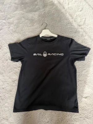 Svart t-shirt från Sail Racing - Svart t-shirt från Sail Racing med vit logga och text på bröstet. Klassisk rund halsringning och korta ärmar. Tillverkad i mjuk bomull för en bekväm känsla. Perfekt till jeans eller shorts för en clean look.
