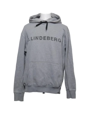 Grå hoodie från J.Lindeberg - Snygg grå hoodie från J.Lindeberg med svart logga framtill. Klassisk modell med huva och svarta dragsnören samt en stor magficka. Mjuk bomullskänsla och ribbade muddar vid ärmslut och nederkant. Perfekt för en avslappnad stil.